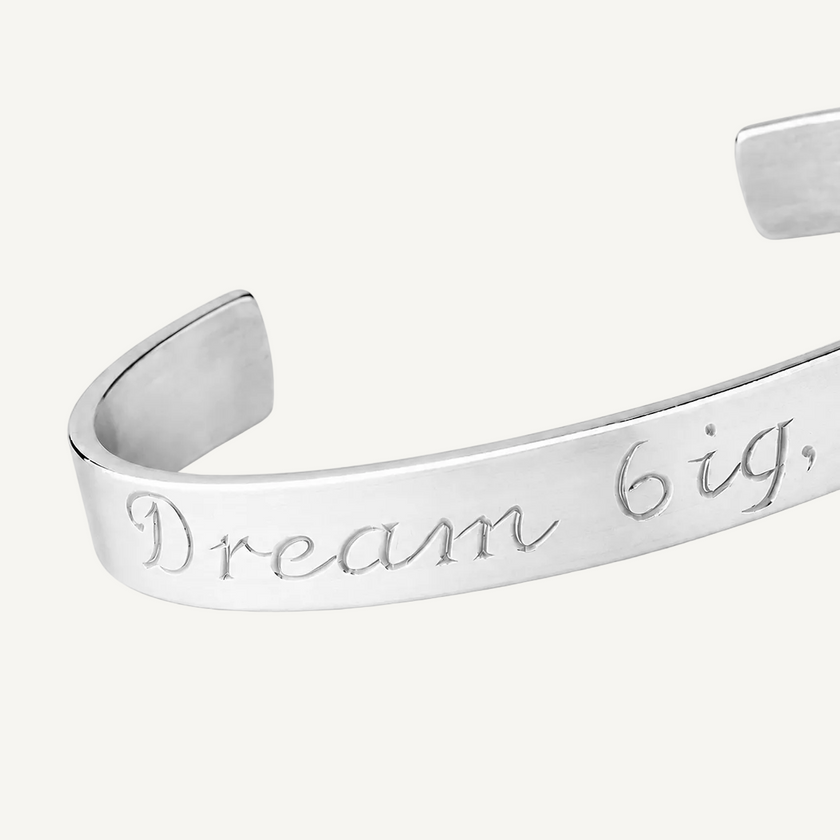 A silver metal cuff bracelet engraved with the phrase 'Dream big.' in a cursive script, shown on a clean white background.