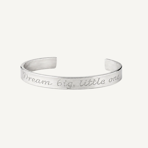 A silver metal cuff bracelet engraved with the inspirational quote 'Dream big, little one' in a cursive font, shown on a clean white background.